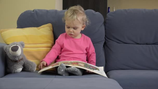 Эмоциональная девочка переворачивает страницы книги, сидя на диване с плюшевым мишкой — стоковое видео