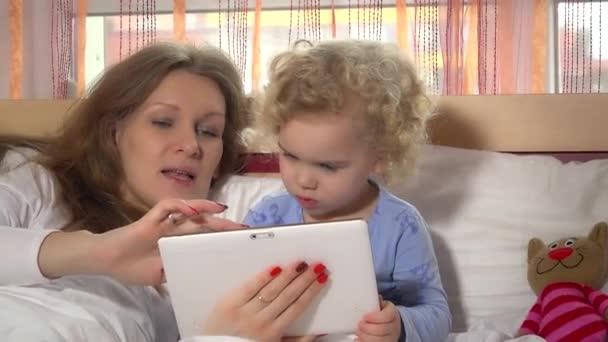 Menina criança curiosa com sua mãe brincando com computador tablet na cama — Vídeo de Stock