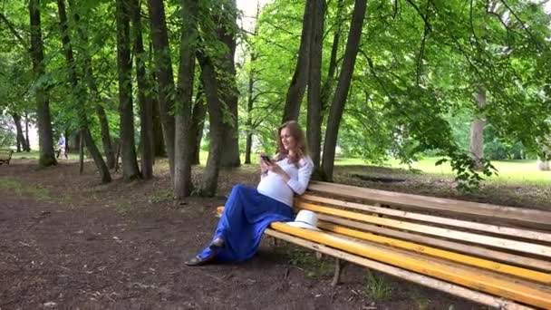 Femme enceinte bavarder sur le téléphone portable tout en étant assis sur le banc du parc à l'extérieur . — Video