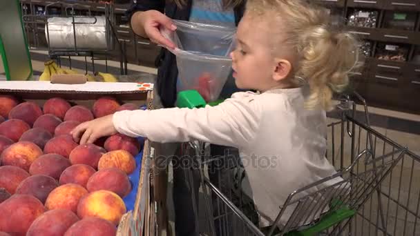 Linda niña en coche de compras ayudar a la madre a recoger frutas de melocotón en el supermercado — Vídeo de stock