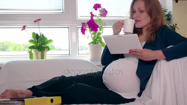 Lekfull gravid affärskvinna dricka kaffe och använda surfplattan på soffan hemma — Stockvideo