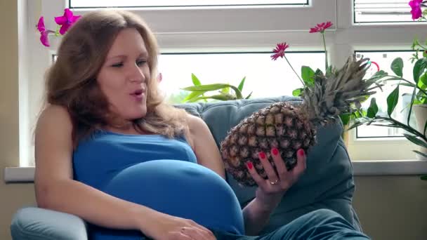 Pregnant woman with big belly holding pineapple ananas fruit sitting on sofa — Stock Video