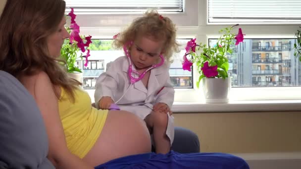 Roztomilá holčička a těhotná matka hrát Doktor s stetoskop sedět na pohovce — Stock video