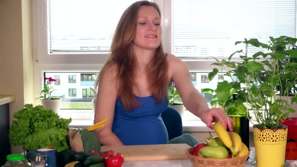 Hongerige glimlachend zwangere vrouw met grote buik afschilferen van bananen in de keuken — Stockvideo