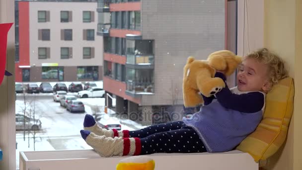 Nettes kleines Mädchen, das mit einem Teddybär auf einem Heizkörper am Fenster liegt. Schneefall im Winter — Stockvideo