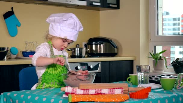 Милая маленькая девочка-повар в белой шляпе смешивает муку для торта на столе на кухне — стоковое видео