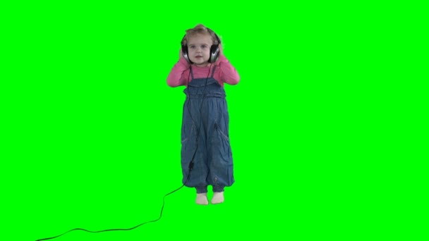 Маленькая девочка с огромными наушниками слушает музыку на зеленом фоне — стоковое видео