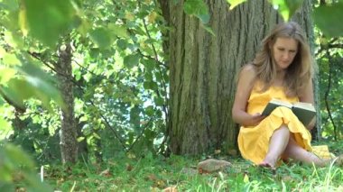 ağacın altında oturup kitap okuma elbiseli güzel kız kitap ve şapka al ve uzaklaş. 4k