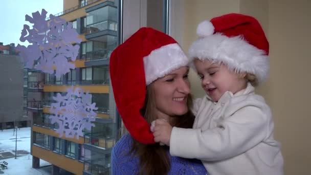 Mor och liten flicka med rött santa hatt ha kul nära fönstret. Snöflingor — Stockvideo