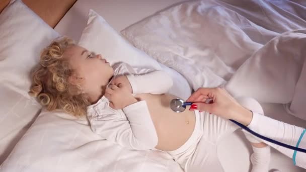 Детский врач осматривает маленькую симпатичную девочку, лежащую в постели со стетоскопом. — стоковое видео