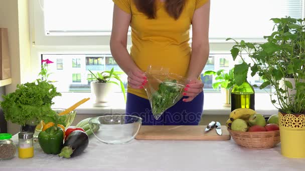 Беременная женщина распаковывает зеленые листья салата и кладет в стеклянную тарелку на кухонный стол — стоковое видео