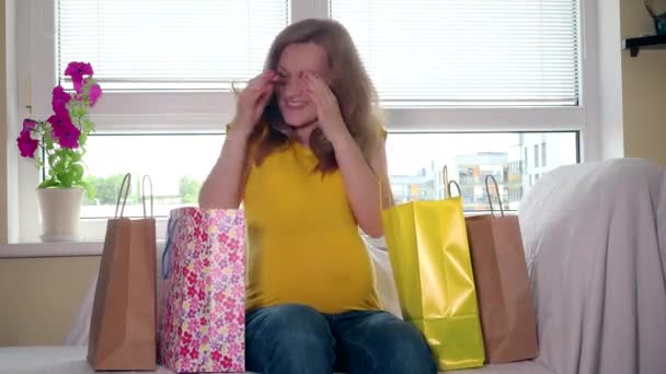 Mujer embarazada feliz con bolsas de colores sentarse en el sofá de vuelta a casa después de ir de compras — Vídeo de stock