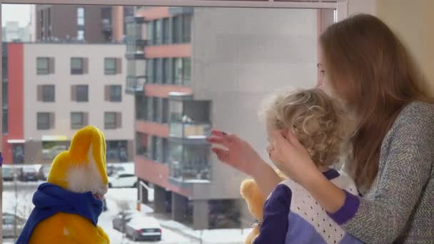 Мама с малышом ребенок девочка сидит у окна и наслаждаться снежинки снег выпадает — стоковое видео
