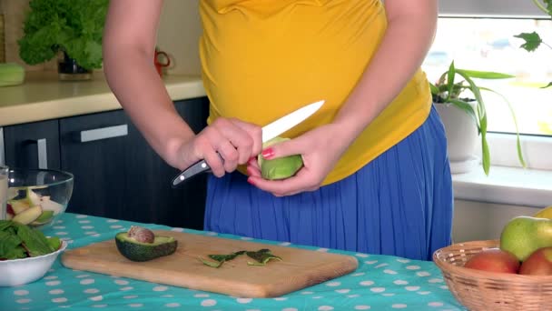 妊娠中の家の妻の腹と自宅の台所でアボカド果実をむく手 — ストック動画