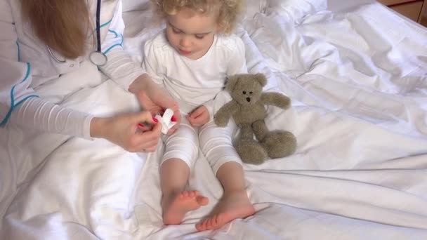 Lekarka ręce trzymać taśmy klejącej na ładny mały dziewczyna nogi w łóżku — Wideo stockowe