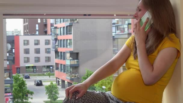 Χαμογελώντας έγκυος γυναίκα μιλά στο smartphone της, κάθεται στο περβάζι του παραθύρου — Αρχείο Βίντεο