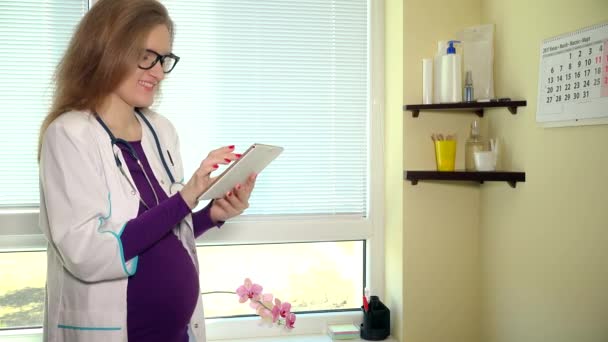 Dokter wanita hamil menggunakan komputer tablet di tempat kerja dan tersenyum melihat ke kamera — Stok Video