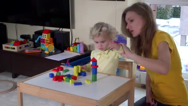 Menina bonito com a mãe jogando tijolos de madeira coloridos perto de pequena mesa em casa — Vídeo de Stock