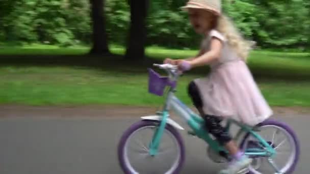 Emocionada niña con el pelo rubio en un vestido rosa monta una bicicleta para niños en la carretera — Vídeos de Stock