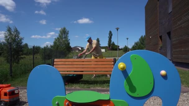 Hombre joven corta césped cerca de los niños colorido parque infantil. Movimiento de cardán — Vídeos de Stock