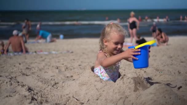 Buried little girl take toy bucket with water from mother and pour it on sand — Stock Video