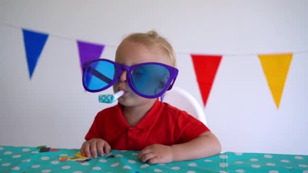 Cute boy with huge glasses and party noisemaker by birthday table. Gimbal motion — Stock Video