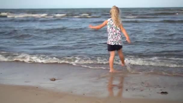 Niño salpicando olas de mar con las piernas. Chica rubia jugar con el agua en la costa — Vídeos de Stock