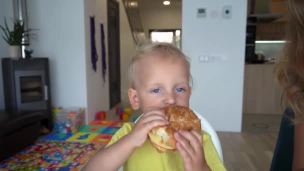 Divertido hijo pequeño niño comiendo hamburguesa sentado junto a la mesa con mamá — Vídeo de stock