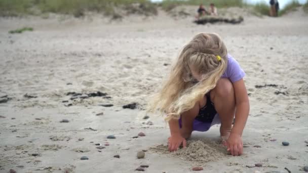 Mooi klein meisje dat met zand aan de kust speelt. Wind verplaatsen mooi blond haar — Stockvideo