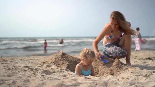 穿着比基尼的妈妈把她的金发小女孩埋在沙滩上的沙子里。 相机运动 — 图库视频影像