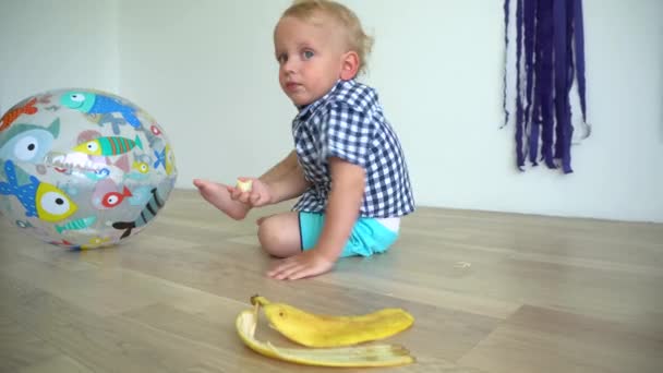 Παιδί δύο ετών κάθεται στο πάτωμα και τρώει κίτρινη μπανάνα. Λαμπερή κίνηση — Αρχείο Βίντεο