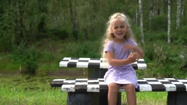 Lustiges kleines Mädchen sitzt auf karierter Bank und lacht im Park — Stockvideo