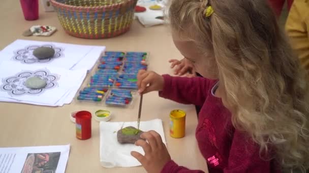 Niña rubia con gouache y pincel pintando piedras de guijarro sentada junto a la mesa — Vídeos de Stock