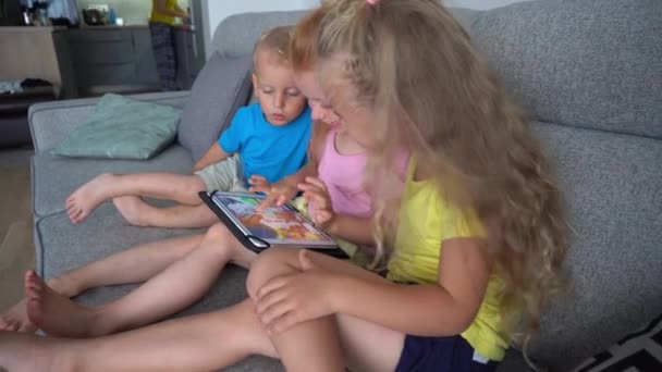 Children using tablet together sitting on the couch. Mother working in kitchen — Stock Video