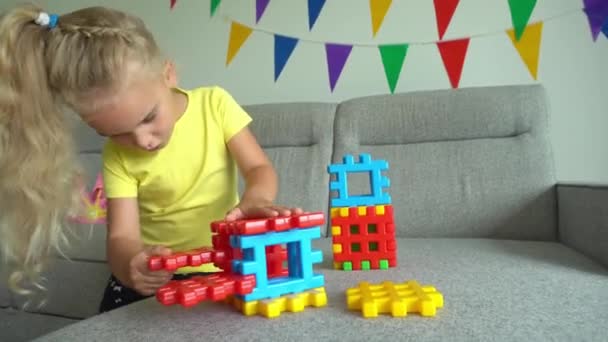 Adorable chica conectando piezas de constructor de colores en el sofá. Movimiento del cardán — Vídeos de Stock