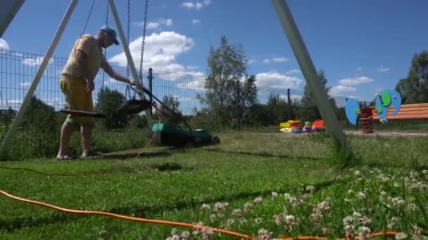 Caucasian man cut grass with lawn mower near swing in playground. Gimbal motion — 비디오