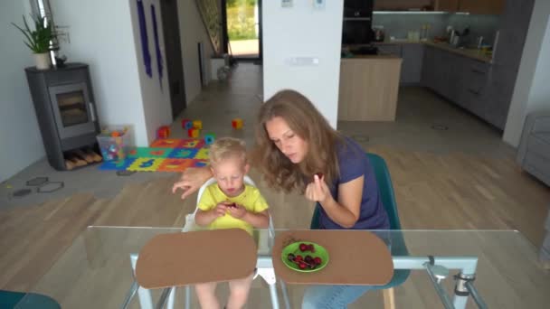 Ξανθό παιδί και καυκάσιος γυναίκα τρώει κεράσι μούρο κάθεται από γυάλινο τραπέζι — Αρχείο Βίντεο