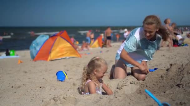 Mooie moeder begraaft haar dochtertje in zand op het strand. Wazige mensen — Stockvideo