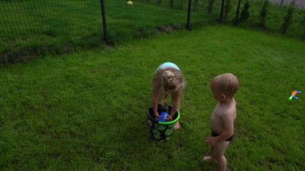 Nettes blondes Schwestermädchen gießt Wasser auf ihren kleinen Bruder Junge aus Spielzeugeimer — Stockvideo