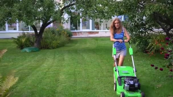 Jovem mulher loira cortando gramado no jardim residencial na noite de verão — Vídeo de Stock