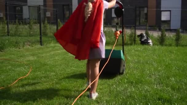 Super girl girl in red cloak mowing lawn in fenced house yard. Corded lawn mower — ストック動画