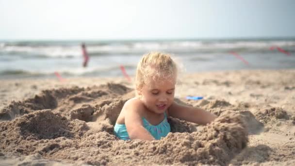 Una niña de 5 años enterrada en la arena se libera y huye. Gente borrosa y mar — Vídeo de stock