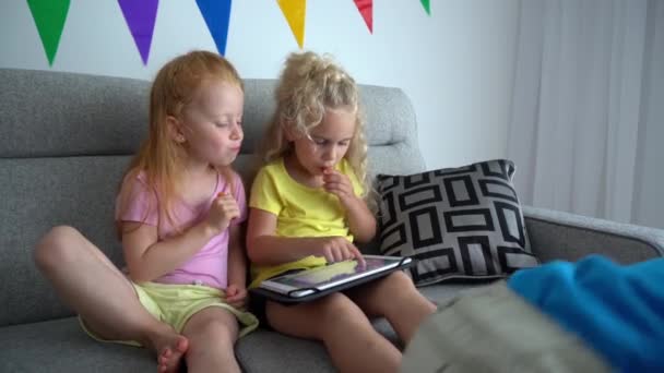 Rödhårig flicka äter morot och blond en med hjälp av tablett. Lillebror sitter på soffan — Stockvideo