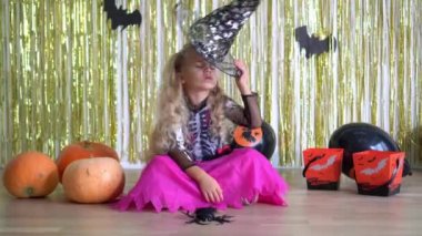 Little angry blond girl with put on witch hat and look at camera near pumpkins