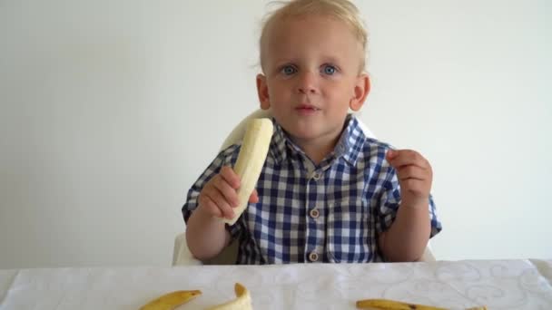 無邪気な顔の子供の白の背景にテーブルの近くに座ってバナナを食べる。ジンバル・モーション — ストック動画