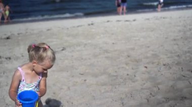 Toddler girl carrying toy bucket with water through beach sand. Blurred people
