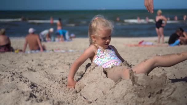Девушка, похороненная в песке, освобождается и веселится с матерью. Размытые люди и море — стоковое видео
