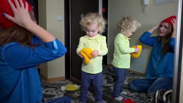 Girl and mother measuring knit hat from big mirror in room. motherhood. 4K — 图库视频影像