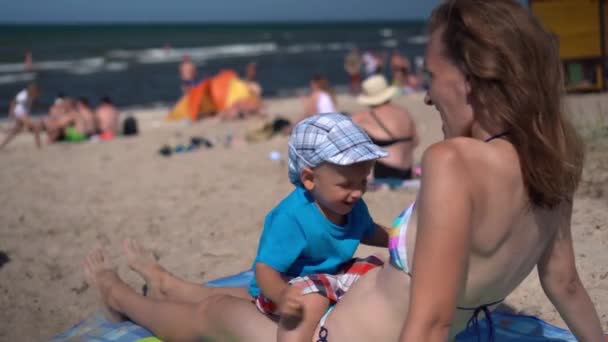 Madre sexual en bikini con hijo pequeño en su estómago acostado en la playa — Vídeos de Stock