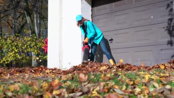 A női munkás leveleket fúj a garázs bejáratáról. Levélfúvót használó lány — Stock videók